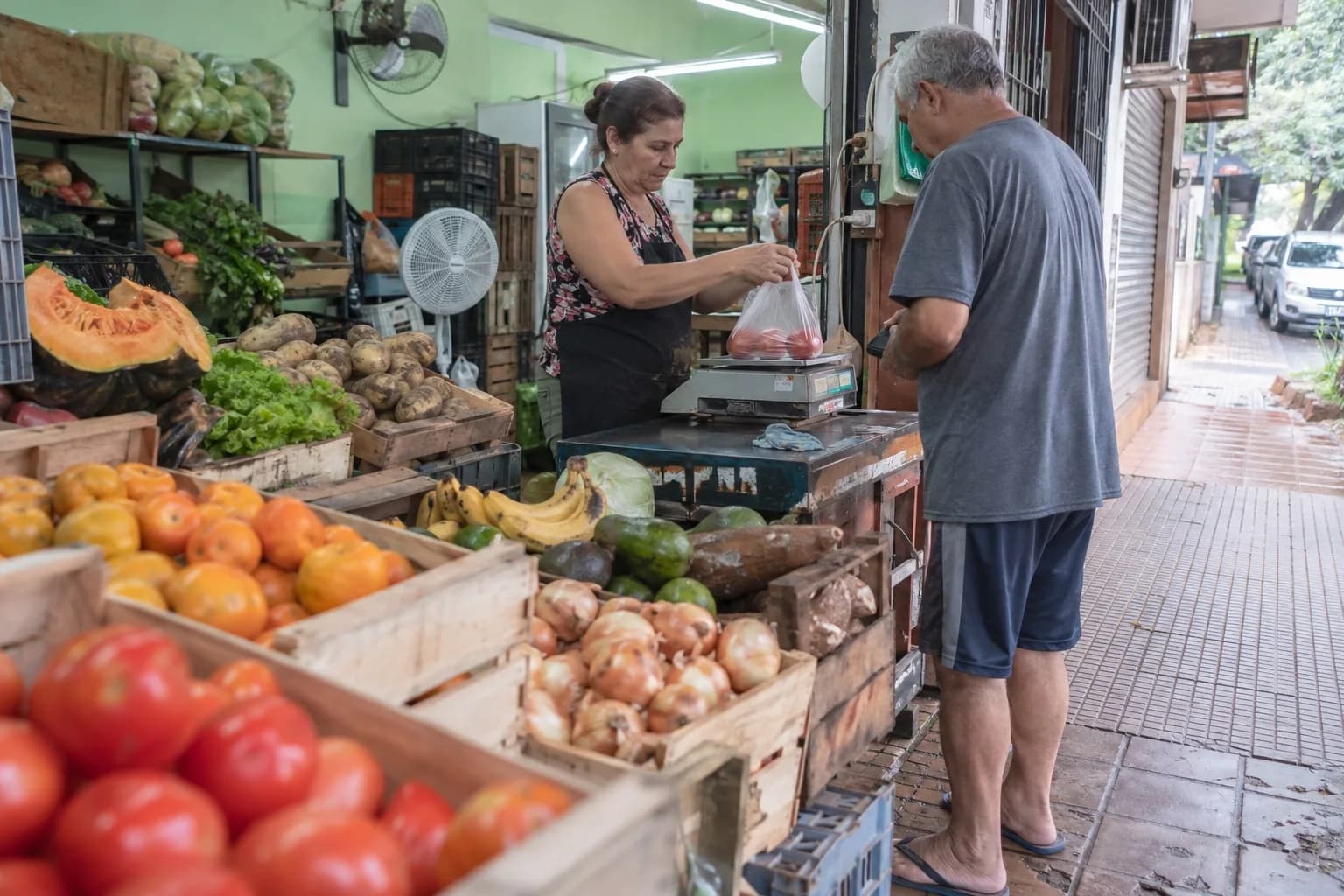 Verdulera pesando tomates para un vecino junto a los cajones de frutas y verduras